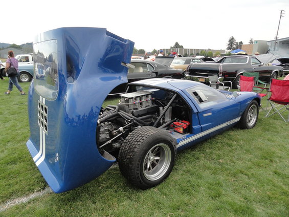 Blue race car with hood open, showing engine and battery. White stripes on hood and side. Other cars and chairs in background