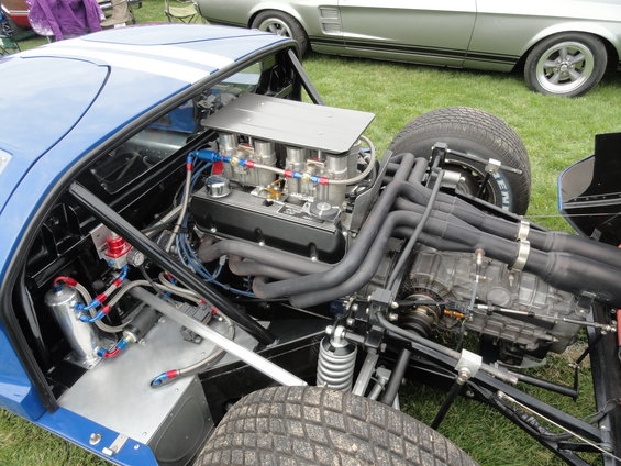 Mid-engine race car with exposed V8 engine, intake manifold, exhaust headers, and suspension components. Another car is visible in the background