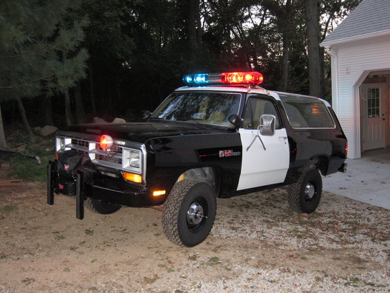 Black and white police SUV with light bar, parked on gravel near a garage