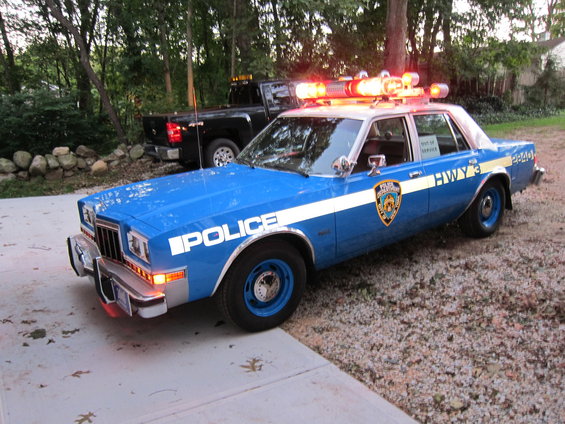 Blue police car with "POLICE" and "HWY 3" markings, parked next to a black pickup truck