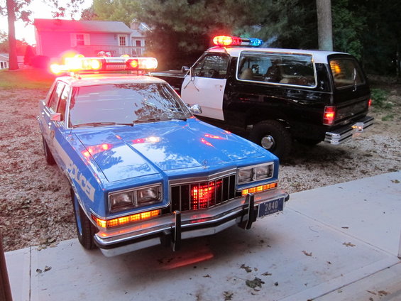Blue and white police car with lights on, parked next to a black and white SUV police car