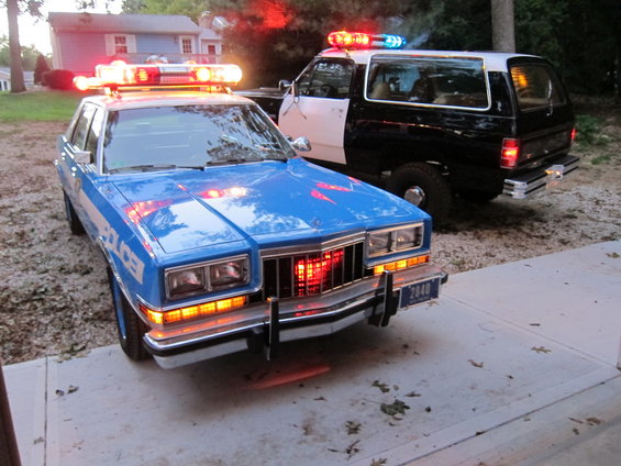 Blue and white police sedan with light bar, parked in front of a black police SUV