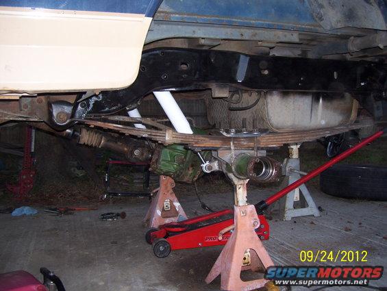 Undercarriage of a vehicle being worked on, supported by jack stands and a floor jack. Leaf springs and fuel tank are visible