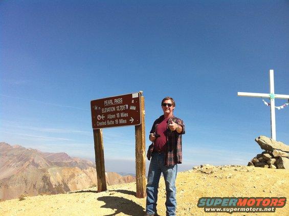 Man in plaid shirt and jeans gives thumbs up near Pearl Pass sign at 12,705 ft elevation