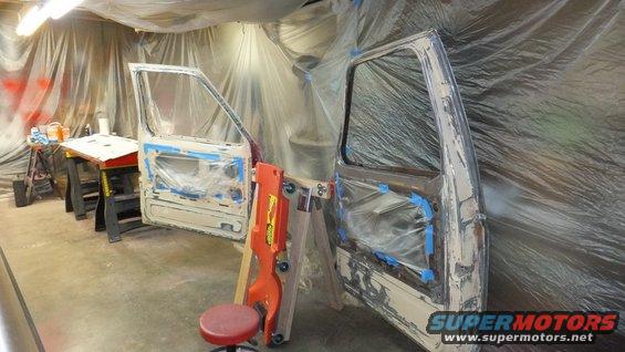 Two vehicle doors, covered in plastic and tape, are propped up in a workshop. One door is on sawhorses, the other is on a stand