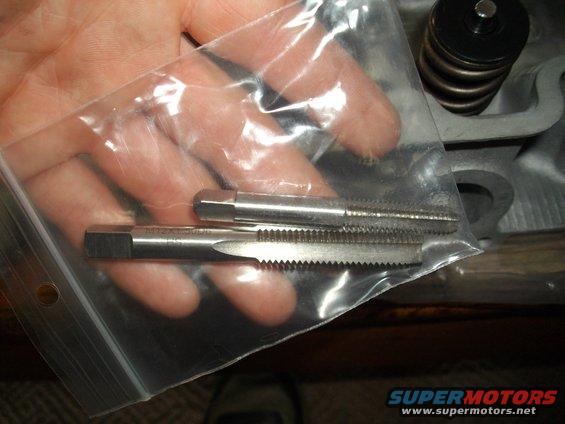 Two metal taps, one with M12x1.50 stamped on it, held in a plastic bag over a metal engine part with a spring