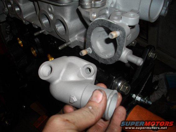 hand holding a gray thermostat housing near an engine block with gasket