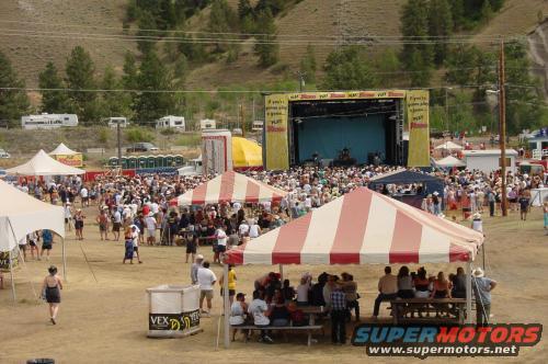 Outdoor festival scene with a stage, large crowd, tents, and RVs in the background