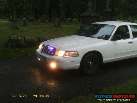 White sedan parked on asphalt in a cemetery with tombstones in background. Headlights and grille lights are on