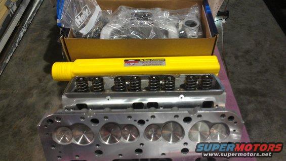 Engine cylinder heads, valve springs, and a yellow bar are laid out on a surface. A box with more parts is in the background