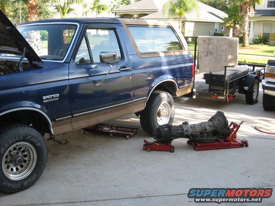 Blue Ford Bronco with hood open, transmission removed, on jacks. Trailer behind