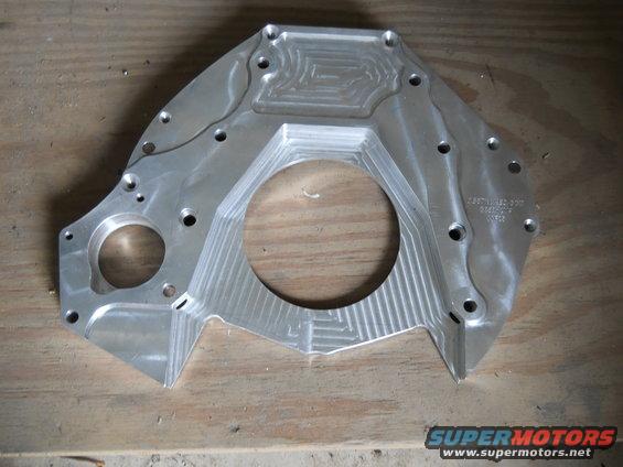 Aluminum adapter plate with machined grooves and bolt holes, resting on a wooden surface