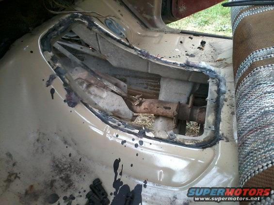 Interior shot of a vehicle's floor pan with a cut-out revealing drivetrain parts underneath. Seat fabric visible on the right