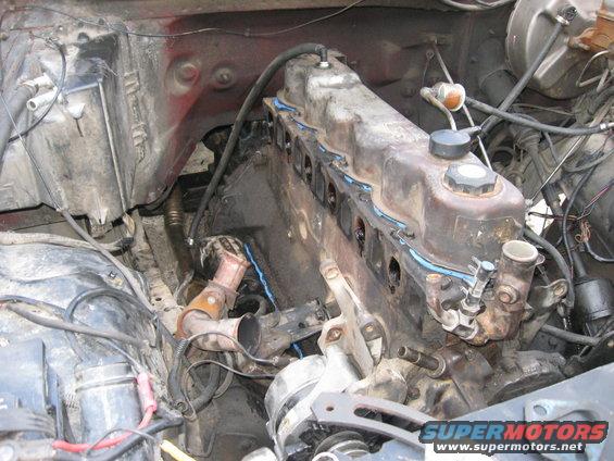 Engine bay showing a V8 engine block, cylinder heads, exhaust manifold, and alternator. Some hoses and wiring are visible