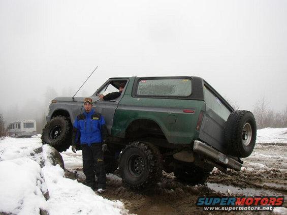 Green Ford Bronco with lifted suspension driving through mud and snow, person standing next to it