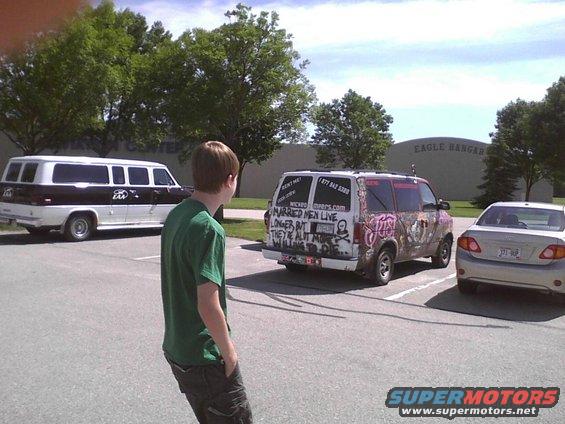 Three vehicles parked in a lot: a long white and brown van, a decorated van, and a silver sedan. A person stands in the foreground