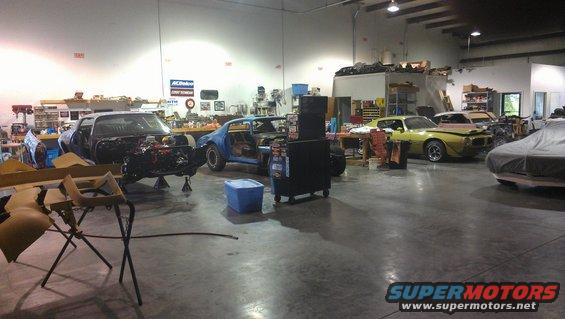 Workshop with three cars in various states of repair; one black car with engine exposed, a blue car with doors removed, and a yellow car with front damage