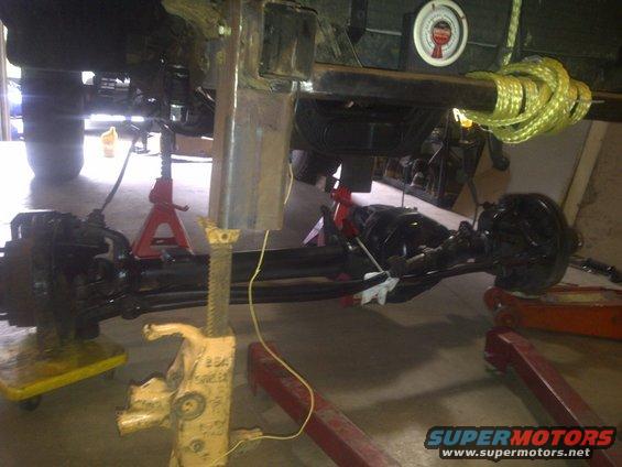 Undercarriage of a vehicle on jack stands, showing suspension and drivetrain components. A gauge is visible on a beam