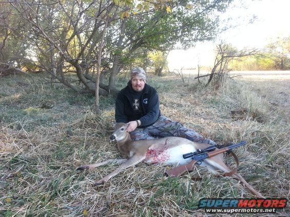 Hunter posing with a harvested deer and rifle in a grassy, wooded area