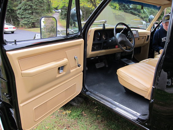 Interior of a truck, showing tan door panel, dashboard, steering wheel, and tan seat. A person is visible in the passenger seat