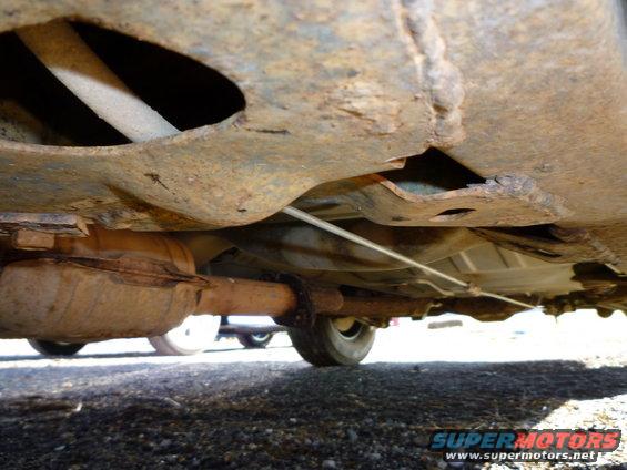 Underside view of rusty exhaust system and frame with a visible tire and another vehicle in the background