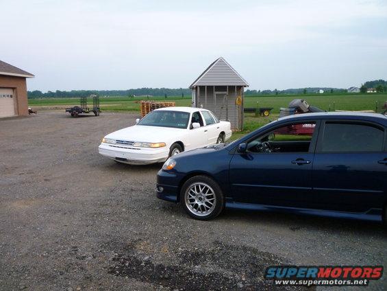 White Ford Crown Victoria and blue sedan parked outside a building with fields in the background