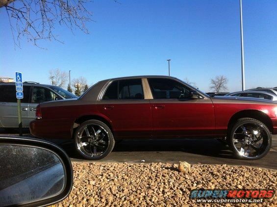 Red and tan 2003 Ford Crown Victoria with large chrome rims parked on gravel