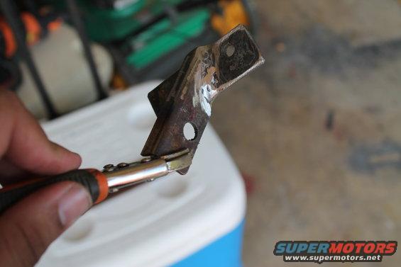 Hand holding pliers on a rusty metal bracket. Background shows a white cooler and garage floor