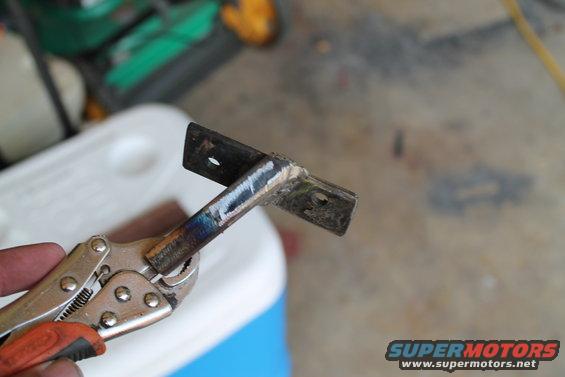 Metal bracket with two holes, held by silver pliers with orange grip. Welded section shows discoloration