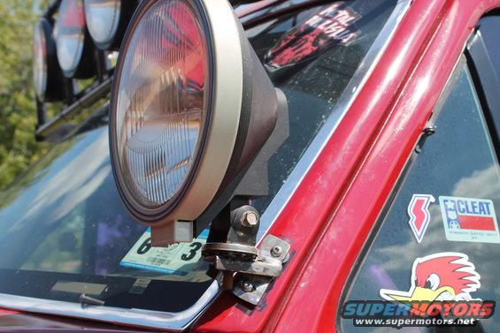 Close-up of an off-road light mounted on the A-pillar of a red vehicle. Stickers are visible on the window