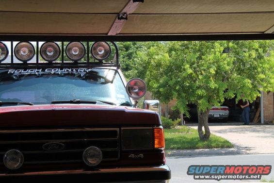Front of red Ford Bronco with roof light bar and 'Rough Country' sticker. Another car is visible in a garage behind it