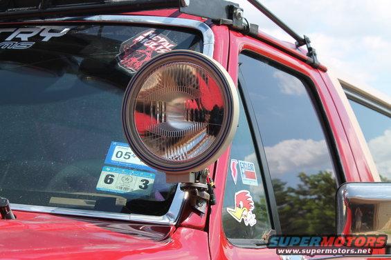 Close-up of an aftermarket auxiliary light on a red vehicle's windshield frame. Stickers are visible on the glass