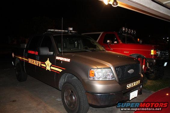 Brown sheriff pickup truck parked next to a red SUV. Text on truck reads "SHERIFF" and "SUPERVISOR"