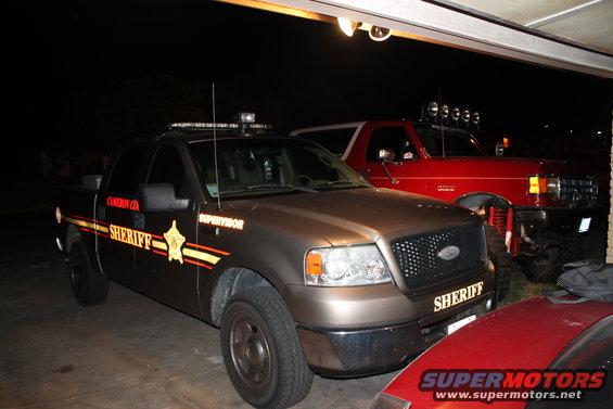 Brown sheriff pickup truck and red Ford Bronco parked in garage. Bronco has auxiliary lights on roof
