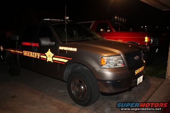 Brown sheriff truck parked next to a red SUV. Sheriff markings on the truck door