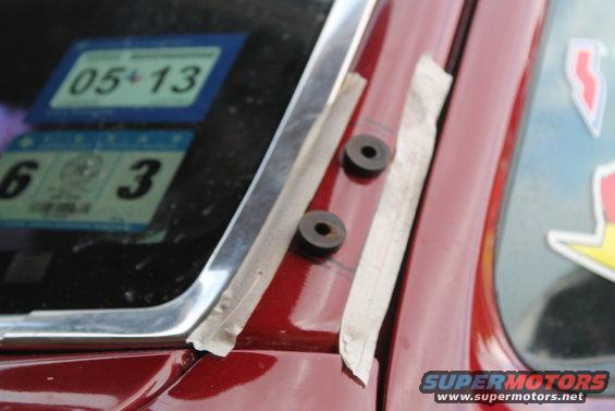 Red car door with two circular rubber grommets taped in place near the windshield frame. Stickers are visible on the glass