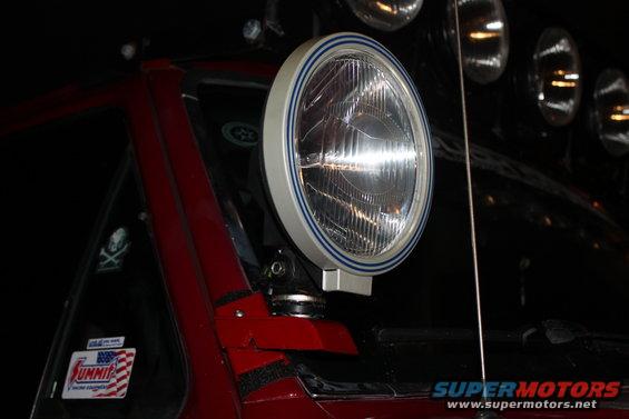 Close-up of an aftermarket auxiliary light with a blue stripe on its bezel, mounted on a red vehicle. A 'Summit' sticker is visible on the window