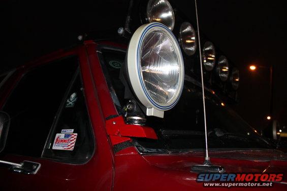 Close-up of a large off-road light mounted on the A-pillar of a red vehicle. Several other lights are visible on the roof