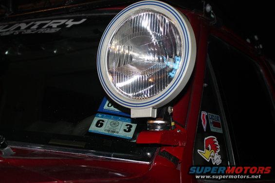 Red vehicle's A-pillar with a large round spotlight attached. Stickers are visible on the glass and body