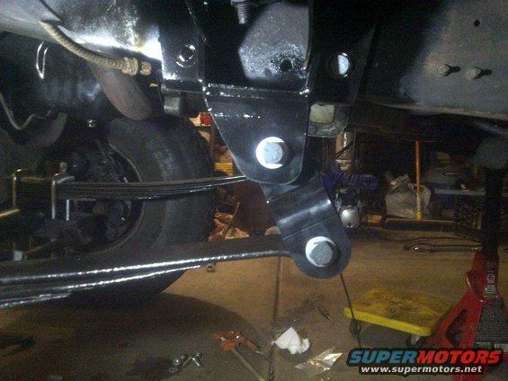 Close-up of a black metal suspension bracket bolted to a vehicle frame, with leaf springs and a tire visible