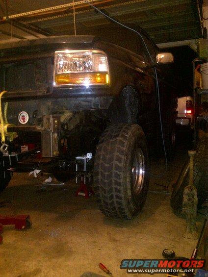 Front end of a dark-colored Ford truck, possibly a Bronco, with the grille removed and front end raised on jacks. Large off-road tires are visible