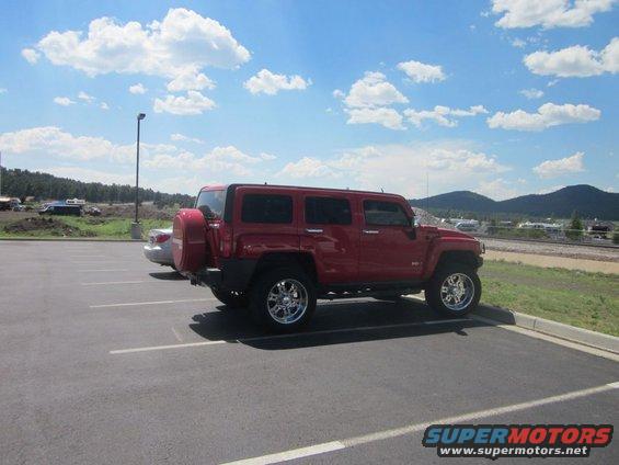 Red SUV parked in a lot, showing its side and rear with a spare tire mounted. Another car is partially visible to the left