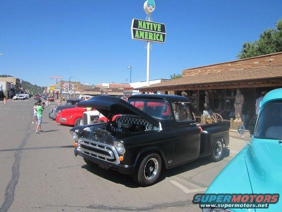 Black 1955 Chevrolet pickup truck, hood open, parked on street with other classic cars. Native America sign visible