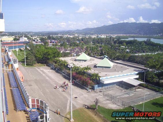 View of a tropical landscape with buildings and a cruise ship, showing a paved area with people and a distant mountain range