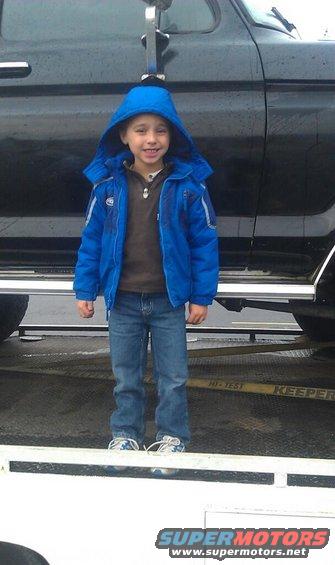 Child in blue jacket and jeans stands on a trailer in front of a black vehicle. Yellow straps are visible