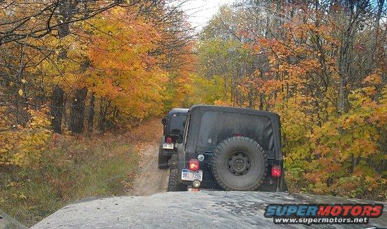 Black Jeep Wrangler with spare tire on rear, driving on dirt road during autumn