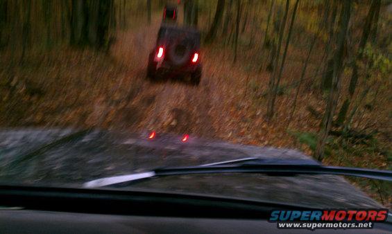 View from inside a vehicle looking at two SUVs driving on a dirt road in a forest during autumn