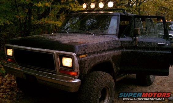 Muddy black Ford Bronco with roof lights, driver's door open. Looks like it's in a wooded area