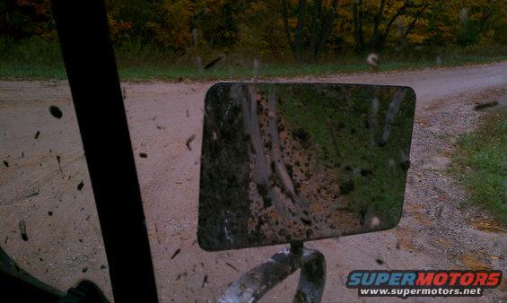 Side mirror reflecting a muddy dirt road with trees in the background