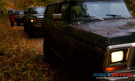 Front corner of a dark 1978 Ford Bronco, followed by other SUVs and trucks on a leaf-strewn dirt road